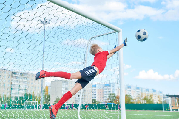 Full length portrait of teenage boy leaping forward and catching ball while defending gate in football match