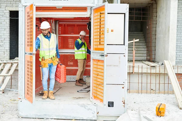 Two men in helmets and waistcoats standing inside industrial lift outside unfinished building