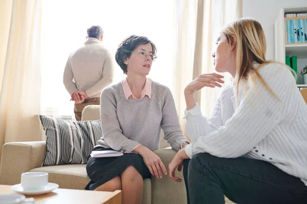 Kısa saçlı ciddi profesyonel psikolog koltukta oturan ve ona tavsiye verirken evli kadının anksiyete dinlerken, kayıtsız kocası arka planda pencerede ayakta