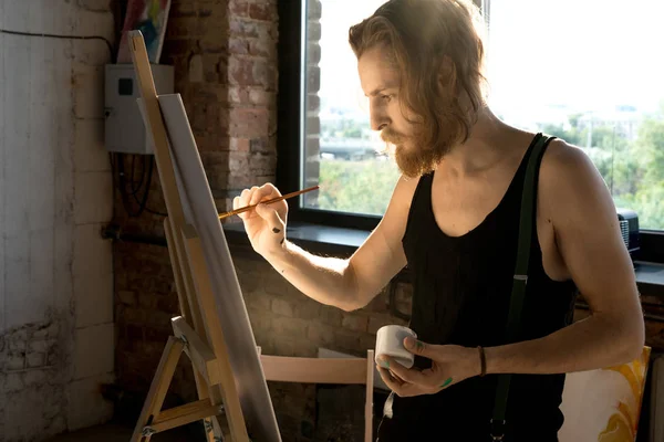 Güneş ışığı ile aydınlatılmış sanat stüdyosunda şelon üzerinde fırça ve resim tutan odaklanmış uzun saçlı sanatçının yan görünüm portresi, kopya alanı