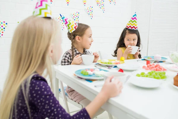 Group of cute multi-ethnic girls in party hats sitting at dining table and drinking tea from cups while chatting at dinner party