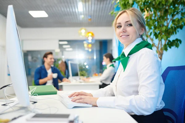 Smiling beautiful young lady wearing white blouse and neck scarf sitting at table and tying on computer keyboard while working in bank and filling online customers card.