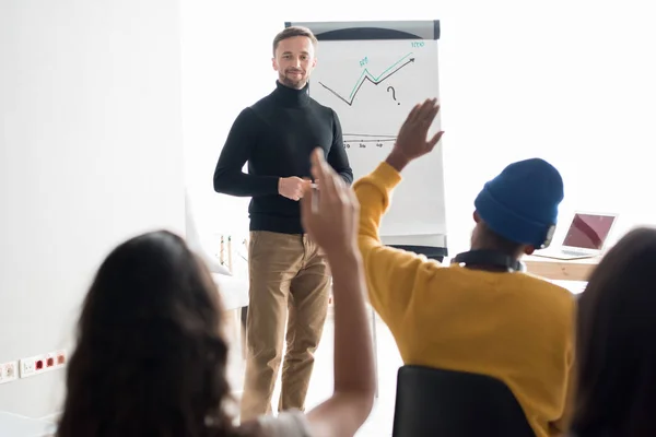 Smiling handsome young bearded speaker in black polo neck standing at whiteboard with graph and answering question of audience in convention center