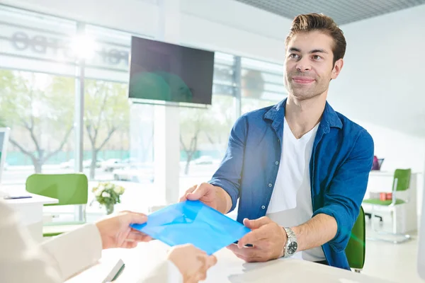 Content handsome young man with stubble sitting at table and taking folder with documents in multifunctional center while getting approved papers