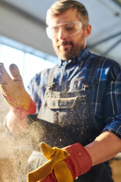 Close-up of serious handsome middle-aged workman in safety goggles dusting off hands while brushing sawdust off work gloves