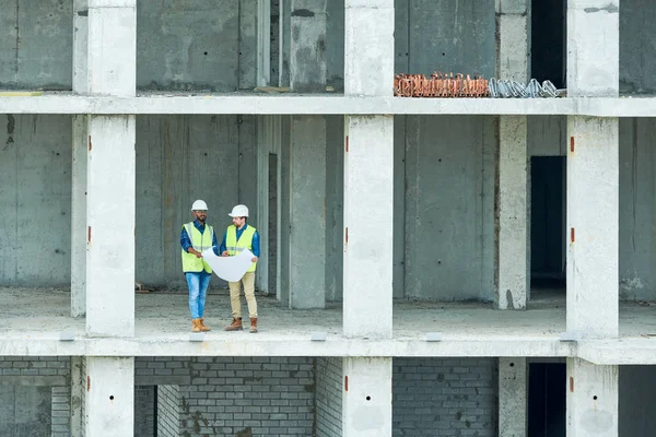 未完成の建物の床に立ち 一緒に建設計画を検討するヘルメットとウエストコートの2人の男性 — ストック写真
