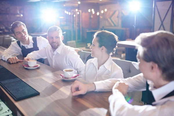Cheerful excited restaurant staff in white shirts sitting at bar counter and drinking espresso while chatting after work