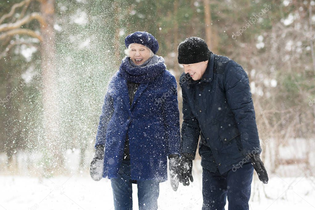 Waist up portrait  of excited senior couple playing with snow in winter forest and laughing happily, copy space