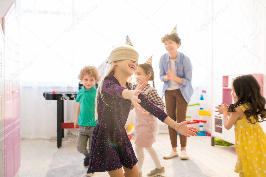Jolly excited girl in eye patch running over room and keeping arms outstretched while catching up with friends during blind mans buff
