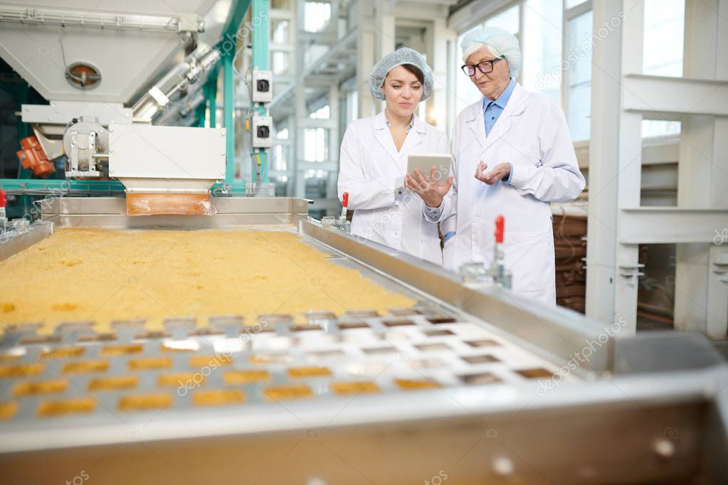Portrait  of  two  women doing  production quality inspection in food factory standing by conveyor belt and using digital tablet, copy space
