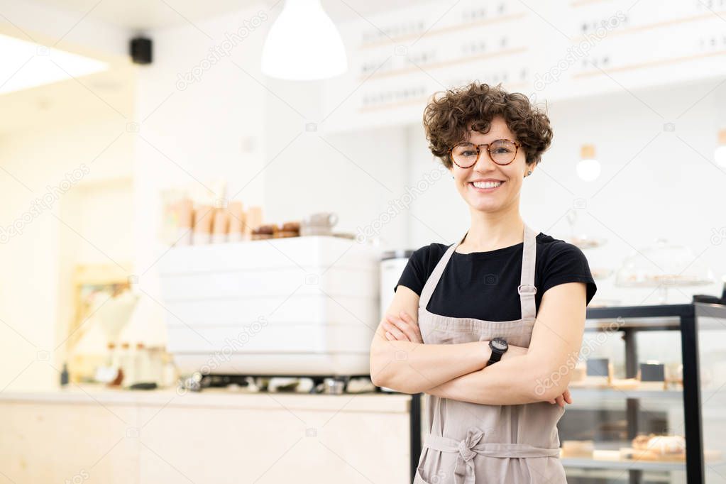 Smiling attractive hipster female barista standing in coffee shop and crossing arms on chest while looking at camera