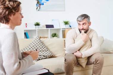 Endişeli, kendine güveni olmayan, rahat bir hırka giyen, koltukta oturan ve terapi seansında profesyonel psikiyatristle konuşurken yastığı ısıran genç bir adam.