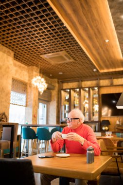 İçerik içe dönük yaşlı adam gözlük ve boğazlı yaka masada oturan ve modern restoranda düşünürken kahve içme