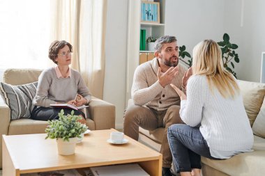 Psikolojik terapi sırasında birbirlerine bağırırken kanepede oturan ve el leri gesturing rahat giyim kızgın evli çift