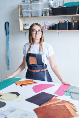 Farklı deri parçaları yla masanın yanında duran ve çalışmaya hazır gözlük takan genç bronzlaştırıcı ustasının portresi