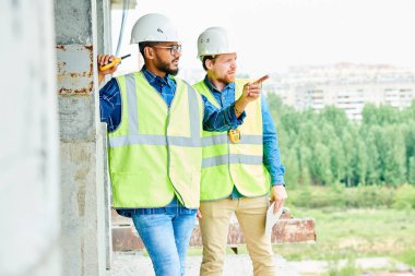 Yeşil yelek ve iş kaskları içinde ciddi dalgın inşaat işçileri yerinde ayakta ve şehirde modern binanın konumunu tartışıyor