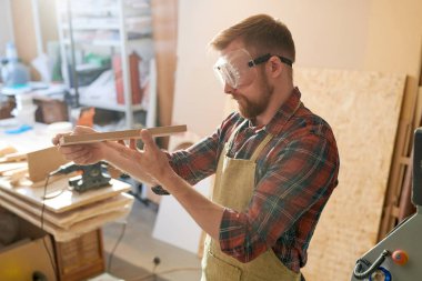 Sakallı marangoz önlük ve güvenlik gözlüğü giyen ve atölyede ahşap detay incelenmesi