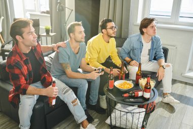 Birgrup genç adam kanepede oturup bira içiyor ve evde birlikte video oyunları oynuyorlar.