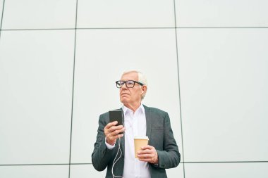 Gözlüklü yaşlı adam sıcak içecek fincan tutan ve akıllı telefon kullanırken ve modern bina duvarının arka planda müzik dinlerken uzağa bakıyor