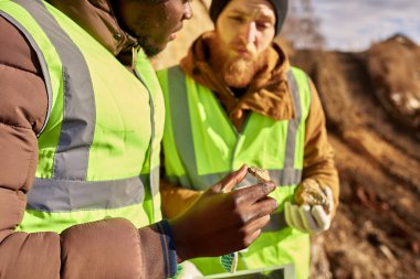 Yansıtıcı ceketler giyen iki sanayi işçilerinin orta bölüm portre, bunlardan biri Afrikalı, yerinde açık havada mineral cevheri teftiş, el tutan taş odaklanmak