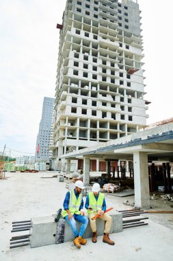 Şantiyede mola sırasında çimento kirişinin üzerinde oturan ve akıllı telefona göz gezdiren kask lı ve yelekli iki adam