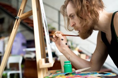 Sanat stüdyosunda şelat üzerine fırça ve resim tutan odaklanmış uzun saçlı sanatçının yan görünüm portresi, kopya alanı