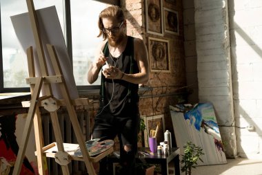 Güneş ışığı ile aydınlatılmış sanat stüdyosunda şöne ile ayakta çağdaş uzun saçlı sanatçı resim resimleri portresi, kopya alanı