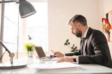 Ofis masasında oturan ve istatistikanaliz ve kağıtları incelerken dizüstü bilgisayar ile çalışan takım elbiseli ciddi meşgul modern işadamı