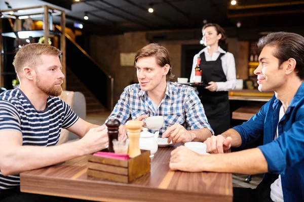 Group of content young friends in casual clothing sitting at table in modern cafe and drinking tea from cups while chatting