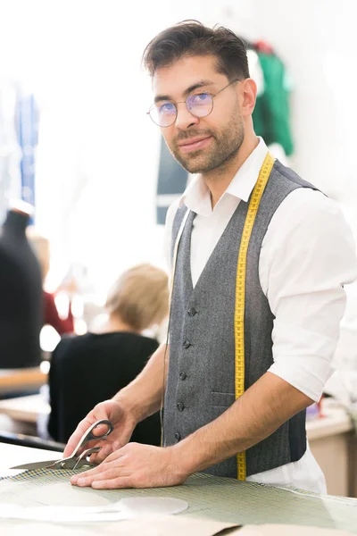 Portrait of smiling male tailor looking at camera and cutting fabric while working in traditional atelier workshop
