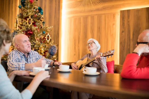 소파에 어쿠스틱 기타를 연주하는 안경을 긍정적 매력적인 할머니는 현대적인 카페에서 — 스톡 사진