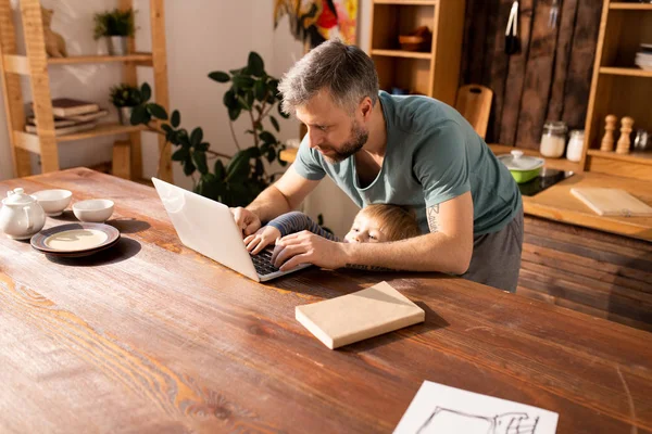 Concentrated handsome bearded father standing at kitchen counter and analyzing information on laptop while little son trying to type on fathers laptop