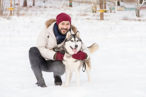 Full length portrait of modern Asian man posing with gorgeous Husky dog sitting down outdoors in winter landscape and smiling happily at camera, copy space