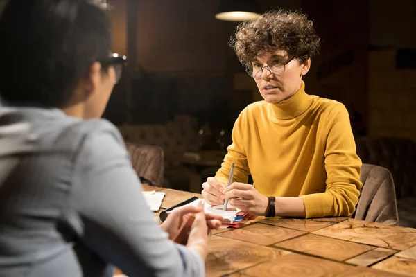 Marketing specialists working late in office: serious curly-haired woman in yellow turtleneck sitting at table with diary and explaining business idea to colleague