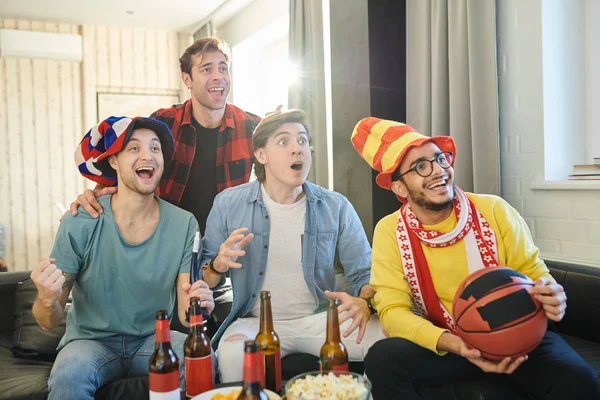 Excited young men sitting in funny hats, drinking beer and watching basketball match on TV at home