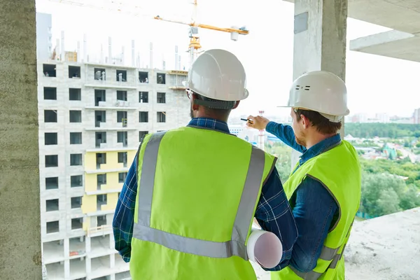 Rear view of constriction workers in hardhats and green vests discussing building structure, male engineer pointing at building under construction