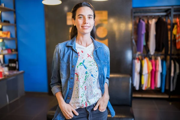 Happy attractive saleswoman in denim shirt and fashionable tshirt with paint splashes holding hands in jeans pockets and looking at camera, she working in youth store