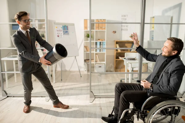 Smiling businessman in suit holding waste basket while his colleague sitting in wheelchair throwing paper in it at office