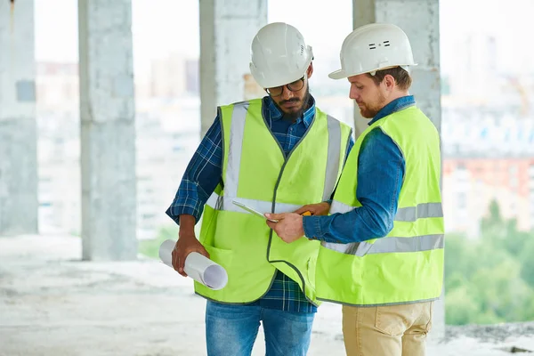 Two Young Men Hardhats Waistcoats Looking Scheme Tablet While Working — Zdjęcie stockowe