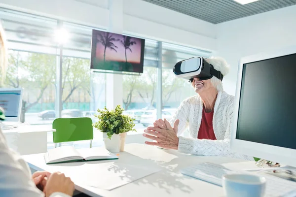 Happy excited senior lady in VR simulator sitting at table with computer and enjoying virtual tour while choosing travel tour in modern agency