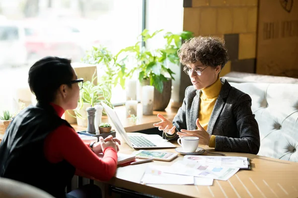 Serious confident female business analyst with curly hair wearing stylish jacket sitting at table in coffee shop and gesturing hands while sharing report results with colleague at informal meeting.