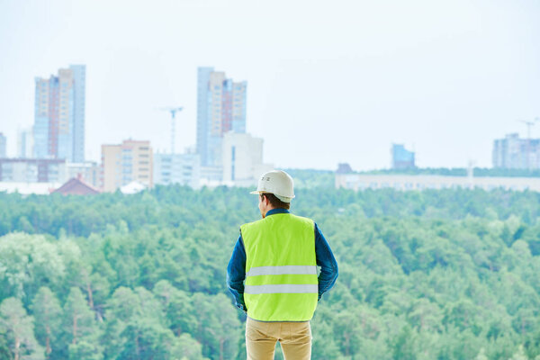 Rear view of male construction engineer in white hardhat and green vest contemplating forests and cityscape while choosing place for new building