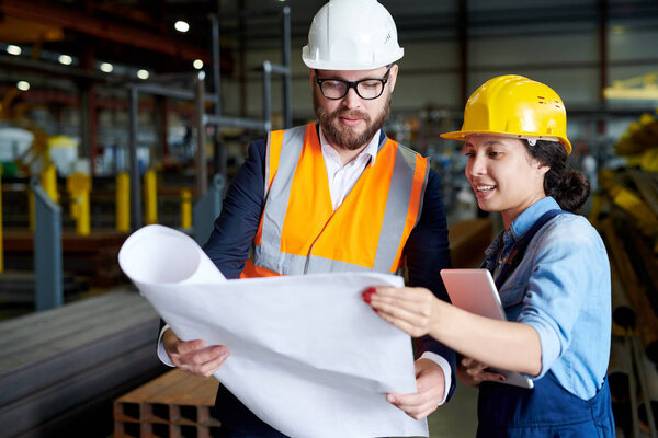 Portrait of modern bearded engineer wearing hardhat holding blueprints  while discussing production with female worker in factory workshop, copy space