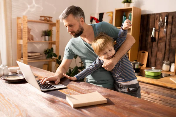 Serious modern handsome father standing at wooden counter and using laptop while trying to work and look after son, little boy getting out of fathers arms