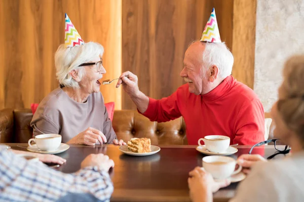 积极细心的老年丈夫在五颜六色的党帽坐在咖啡店的桌子和喂妻子用勺子 — 图库照片