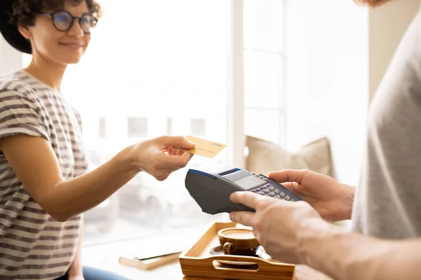 Close-up of smiling attractive young lady giving credit card to cashier while paying for bill in coffee shop