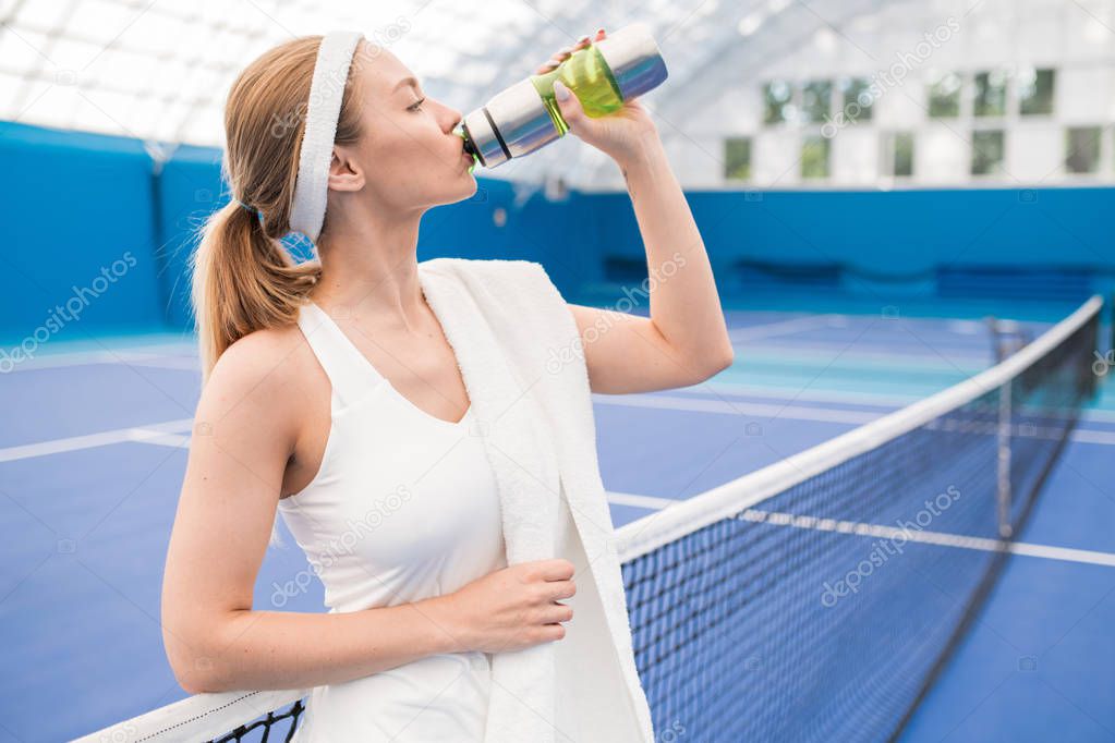 Waist up portrait of beautiful blonde woman drinking water after tennis practice in indoor court, copy space