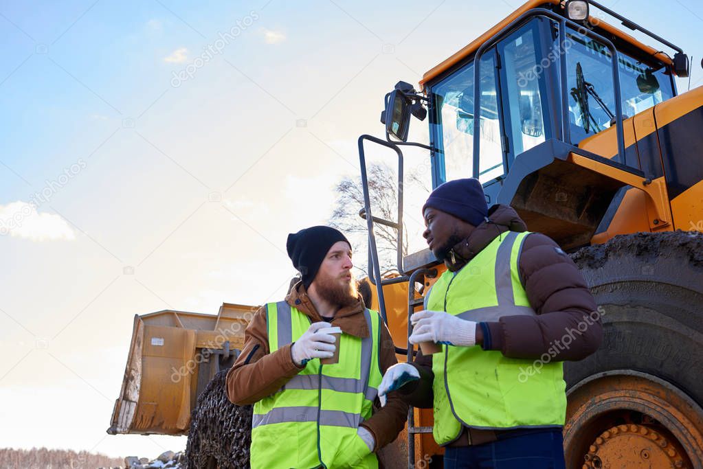 Waist up portrait of two industrial workers, one African-American, drinking coffee and chatting next to heavy industrial truck on worksite, copy space