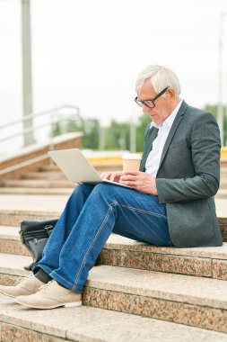Sokak merdivenlerinde otururken sıcak içecek fincan tutan ve modern dizüstü tarama kıdemli adam Yan görünümü