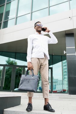 Neşeli yakışıklı genç Afrikalı-Amerikalı işadamı şık giyim modern binaya karşı duran ve cep telefonu ile konuşan, hareket halindeki hayat